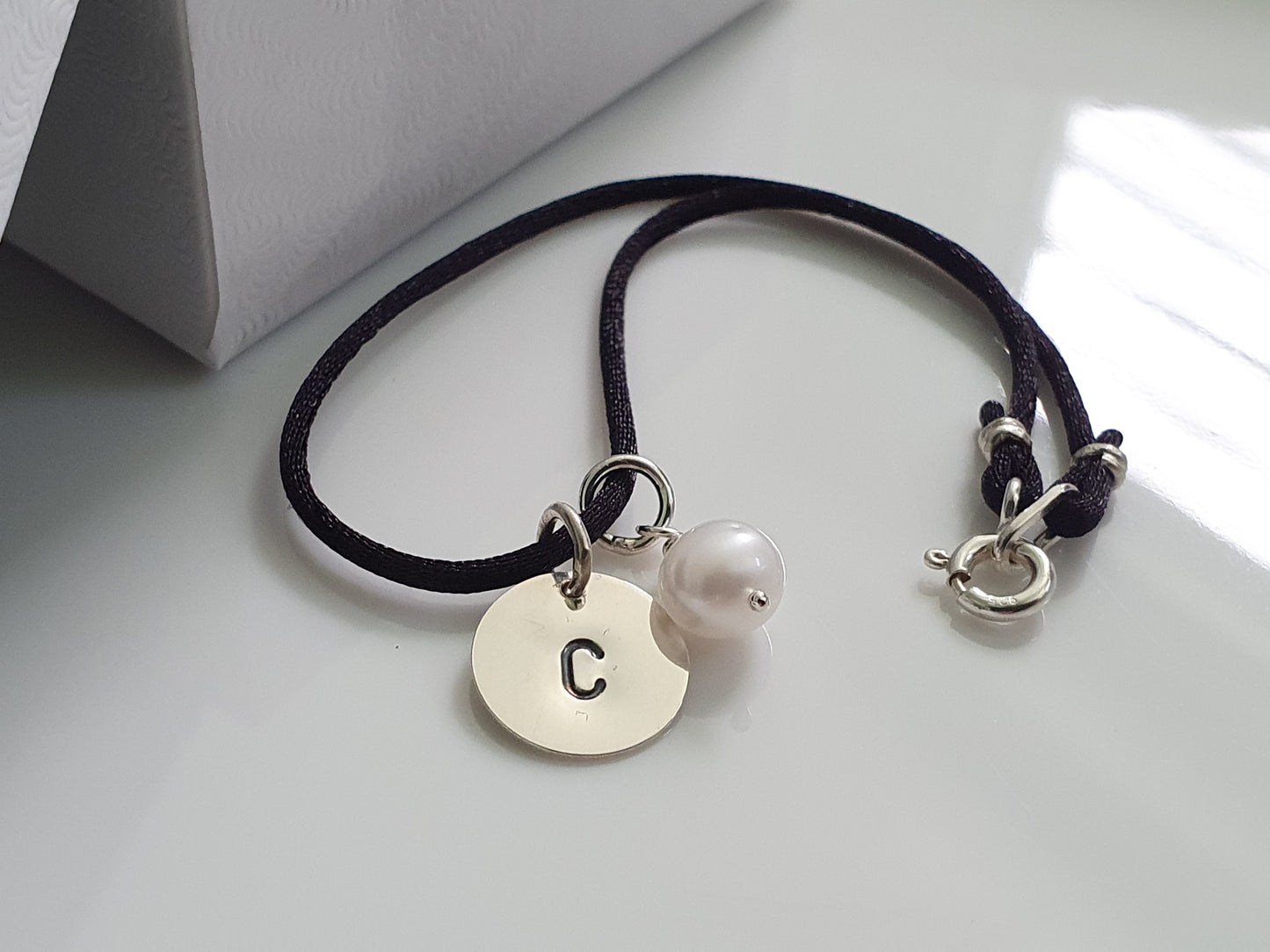 A sterling silver initial charm with a small pearl charm attached to a black satin cord bracelet.