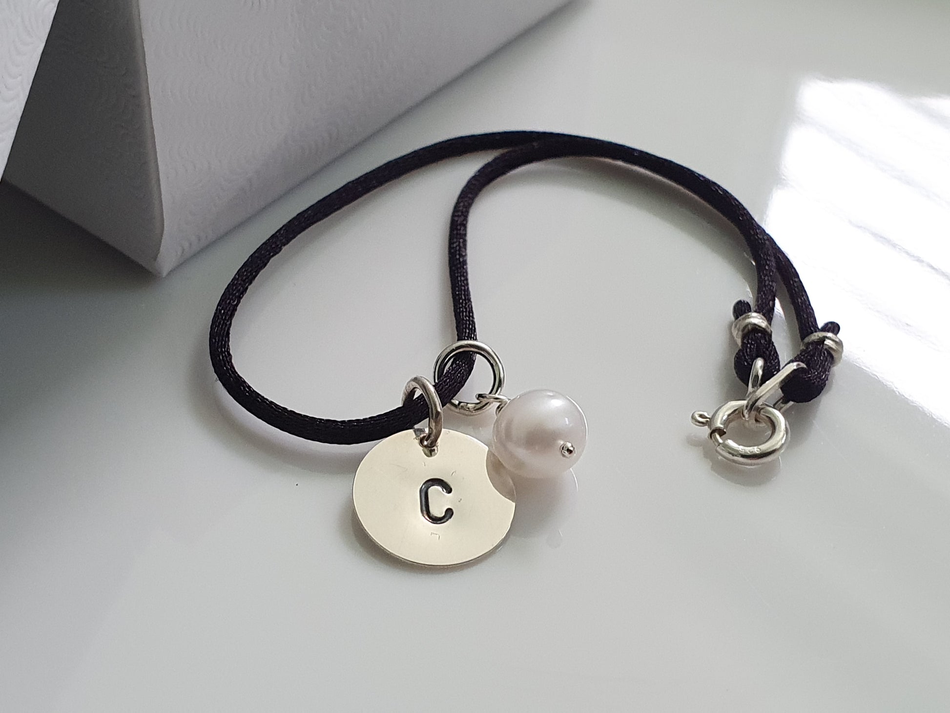 A sterling silver initial charm with a small pearl charm attached to a black satin cord bracelet.