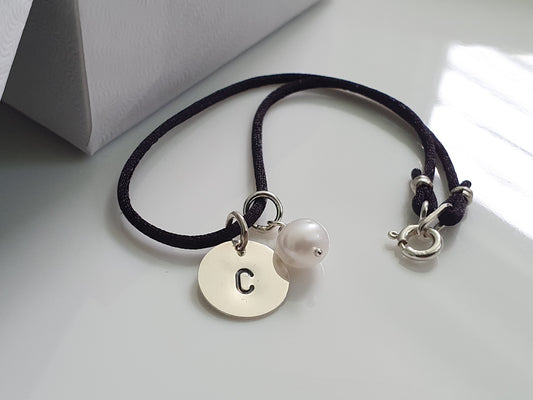 A sterling silver initial charm with a small pearl charm attached to a black satin cord bracelet.